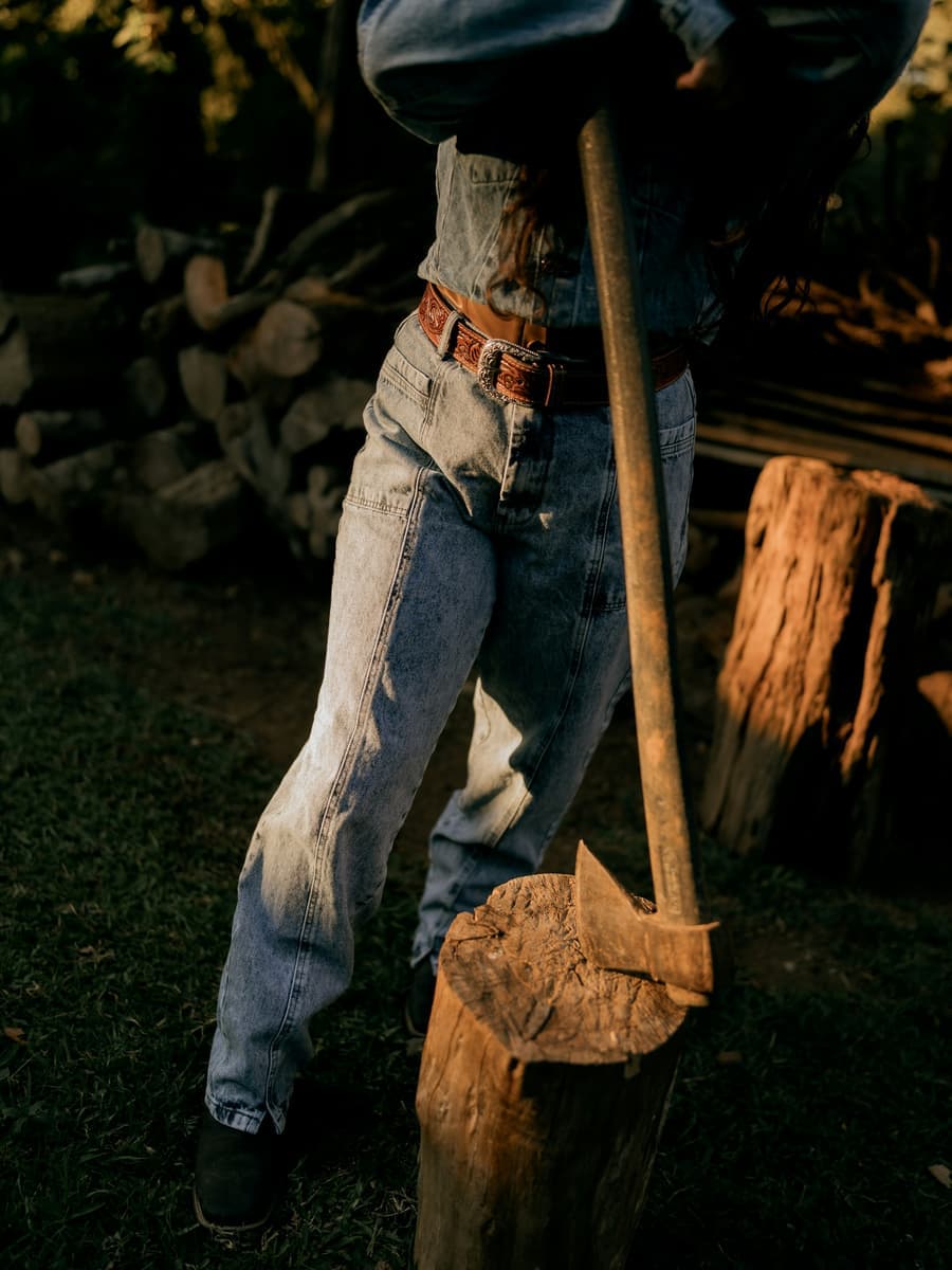 A person in overalls resting an axe on a chopping block in front of a woodpile